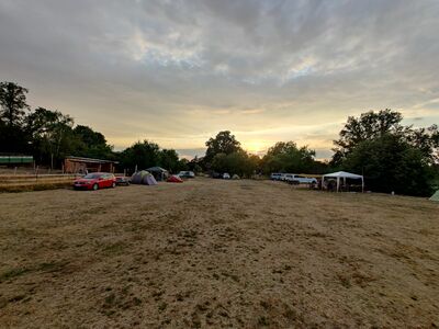 Der Zeltplatz aus Richtung Wald. Die Sonne geht gerade unter, am Himmel sind einige Wolken zu sehen.