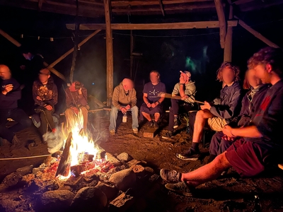 Viele Personen sitzen um ein brennendes Lagerfeuer. Das zum Dreieck aufgebaute Holz brennt hell. Einige Personen haben Stöcke in der Hand, mit deren Hilfe sie Marshmallows ins Feuer halten.