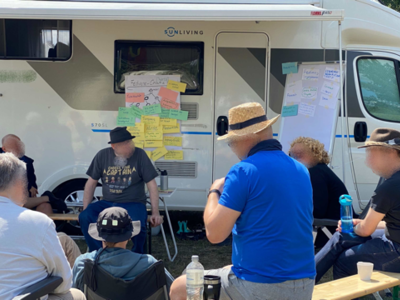Personen sitzen draußen vor einem Camper und spreche miteinander. An der Seite sind Pinnwände mit angehefteten Zettel.