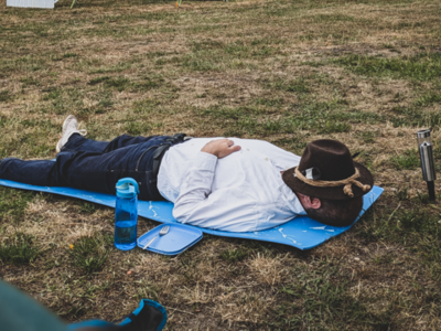 Nach weiter Anreise und langer Nacht kann man schon mal unter der Sonne die Augen schließen. Person liegt auf der Wiese, Hut ins Gesicht gezogen und schläft.