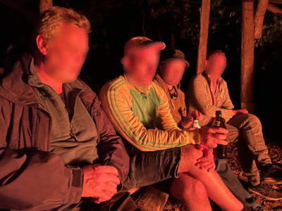 Vier Personen sitzen am Feuer, die Gesichter sind vom Feuerschein rörtlich gefärbt.
