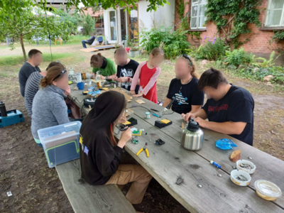 Personen sitzen draußen an einem Tisch und hantieren mit Lötkolben und Zubehör.