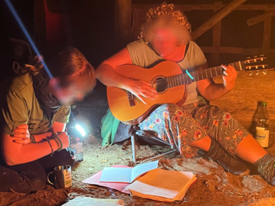 Zwei Sängerinnen sitzen mit Gitarre auf dem Boden vor dem Lagerfeuer.