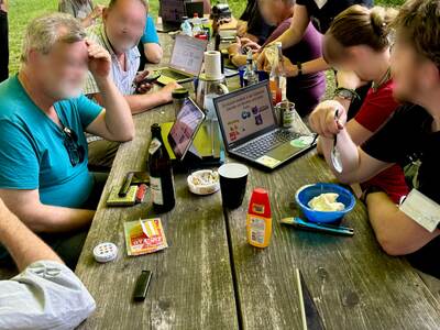 Viele Personen sitzen am Tisch vor dem Gebäude und lauschen einem Vortrag. Die Präsentation wird auf vielen Bildschirmen gestreamt.