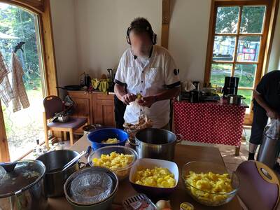 Eine Person mit On-Ear-Kopfhörern baut den Kartoffelsalat zusammen. Auf dem Tisch stehen drei große Schüsseln mit geschnittenen Kartoffeln.