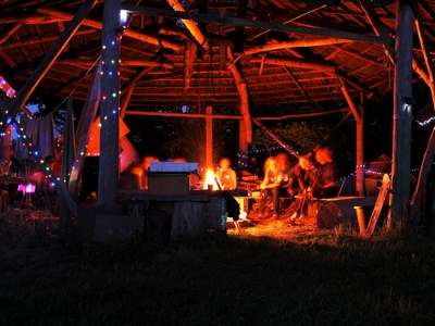 Personen sitzen am Lagerfeuer unter einer Holzüberdachung.