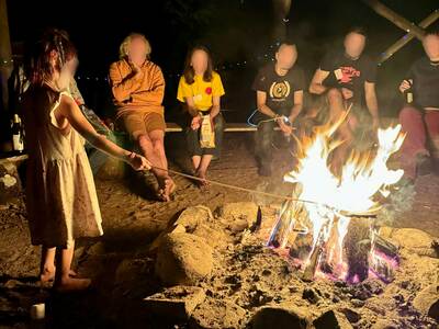Personen sitzen am Lagerfeuer und unterhalten sich. Ein Kind hält einen Stock ins brennende Feuer.