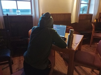 Eine Person mit Kapuzenpullover sitzt im dunkeln am Laptop, man sieht Programmiercode auf dem Screen.
